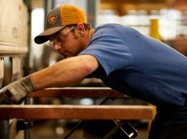 A guy wearing an orange and grey Ariat hat and blue Ariat Rebar Short Sleeved Shirt putting metal into a machine in a workshop