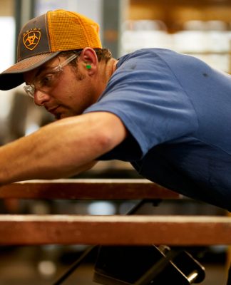 A guy wearing an orange and grey Ariat hat and blue Ariat Rebar Short Sleeved Shirt putting metal into a machine in a workshop