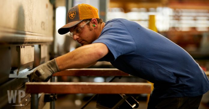 A guy wearing an orange and grey Ariat hat and blue Ariat Rebar Short Sleeved Shirt putting metal into a machine in a workshop